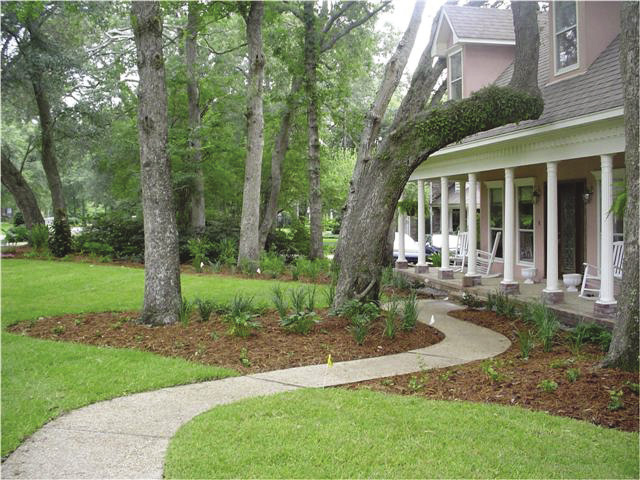 A curved concrete pathway leading to a house. The path is now surrounded by red mulch with several green plants and bright green grass. The clean beds and bed lines surrounding the path and the trees around it create a finished appearance.